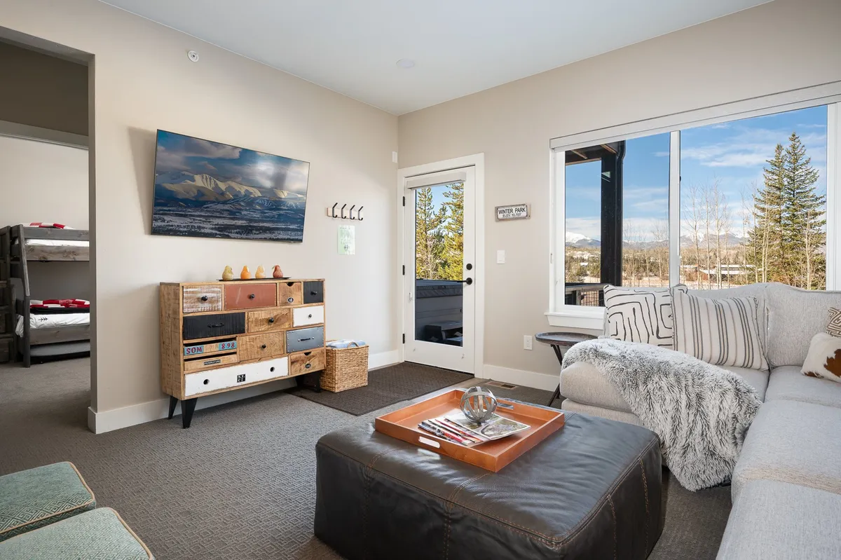 A spacious living room filled with natural light thanks to expansive windows that offer breathtaking views of the surrounding mountains. The room features a cozy sectional sofa, a decorative storage cabinet, and a wall-mounted HD smart TV, creating an inviting atmosphere for relaxation and family gatherings.