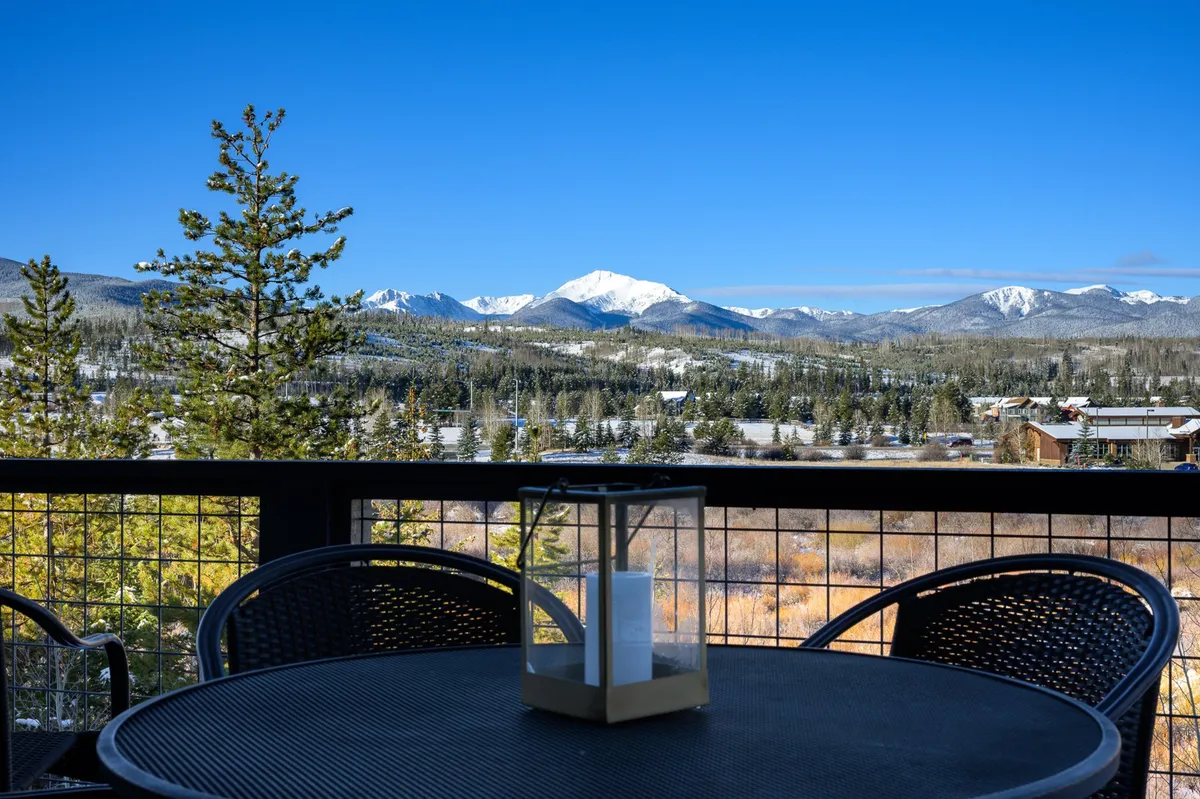 A tranquil balcony scene featuring a table with a lantern, surrounded by lush trees and a dramatic mountain range in the background. The setting invites relaxation and offers sweeping mountain vistas ideal for unwinding or sharing a meal outdoors.