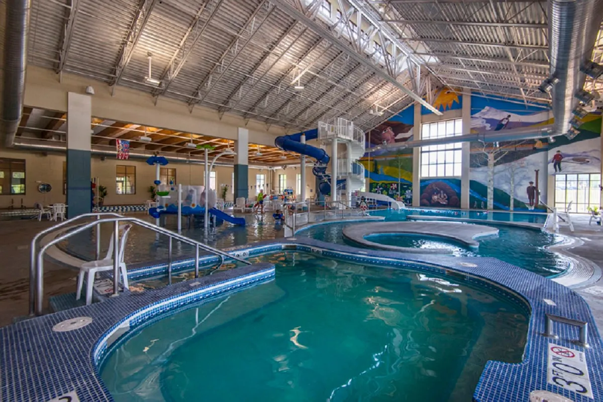 A vibrant indoor pool scene featuring a large swimming pool with a curving water slide, children joyfully playing in the water, and colorful murals decorating the walls. The space is filled with natural light, creating a lively and energetic atmosphere ideal for family fun.