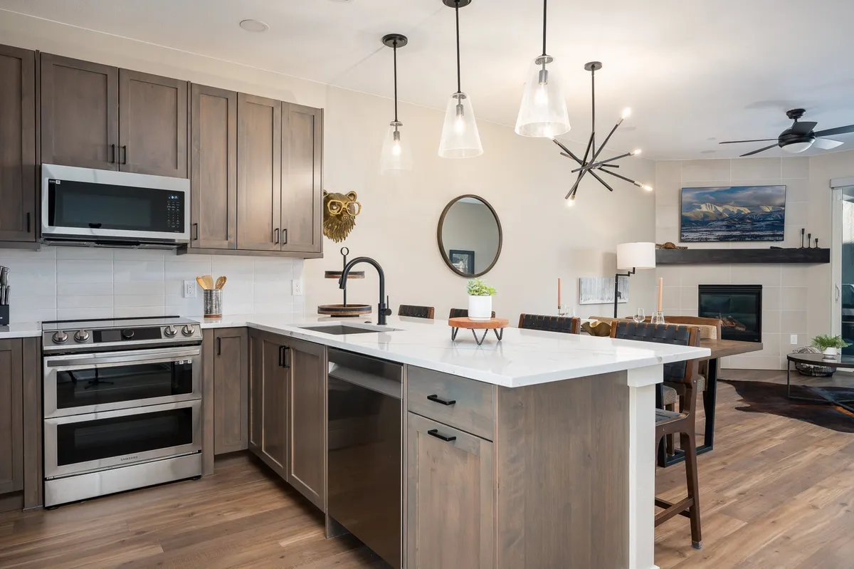 A bright and stylish open-plan kitchen and dining space, featuring a large island with bar stools, stainless steel appliances, pendant lighting, a decorative owl, a round mirror, and a dining area set for a meal. The kitchen is equipped with modern amenities and ample countertop space, seamlessly connecting to the dining area for easy entertaining.