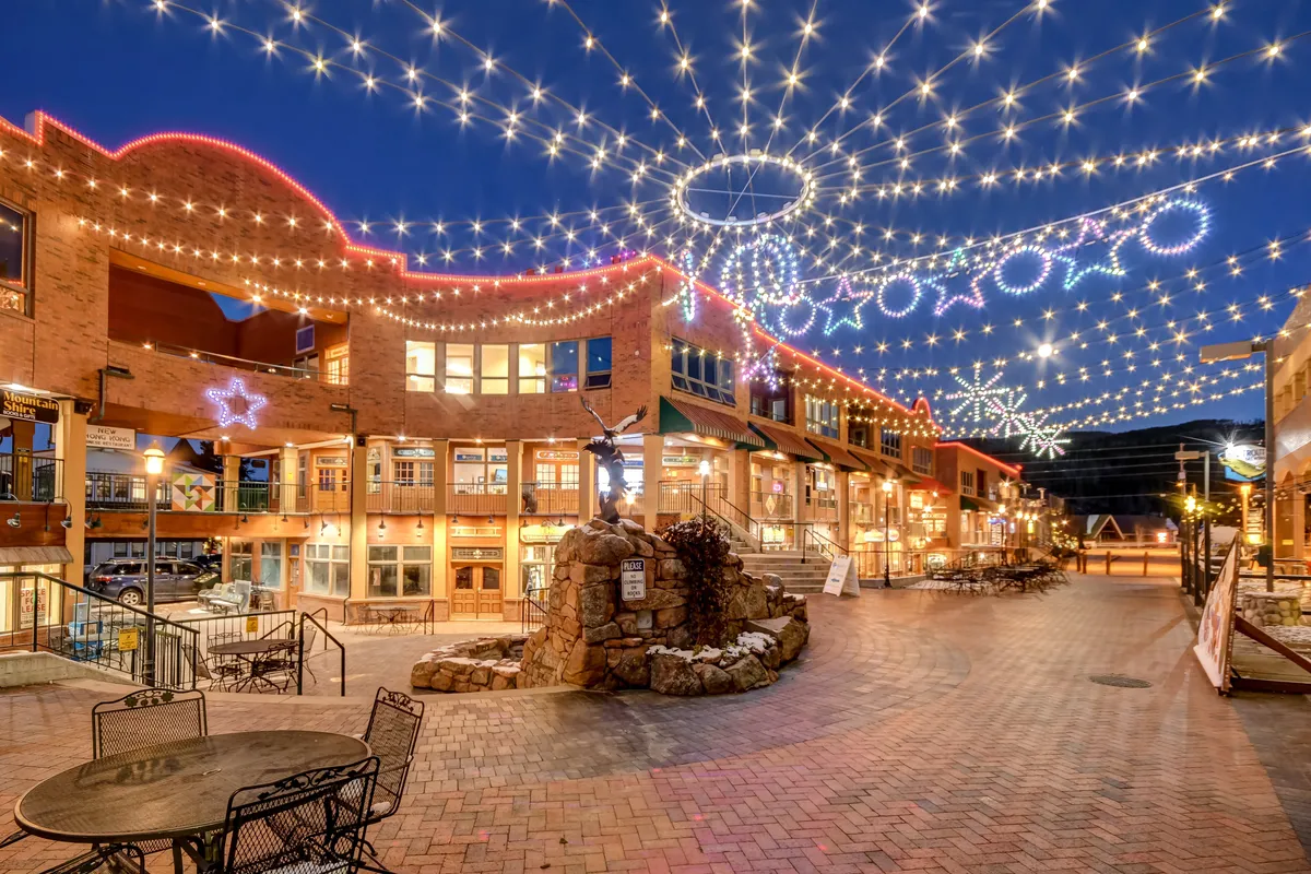 A lively downtown outdoor scene adorned with twinkling string lights and decorative stars. People are dining outdoors, strolling, shopping, and gathering around a festive stone fountain, suggesting a vibrant, year-round community atmosphere.