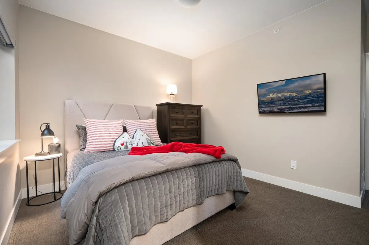 A cozy queen bedroom featuring a bed with a quilted grey cover, red and white striped pillows, a warm red throw, bedside lamp, and framed landscape artwork. Sunlight streams in from a window, adding brightness and warmth to the space.