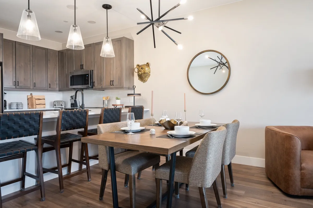 A warm and contemporary dining area adjacent to the kitchen, featuring a dining table that comfortably seats eight, bar stools at the breakfast island, sleek kitchen cabinetry, a modern light fixture, and a decorative sunburst mirror. The ambiance is perfect for both festive gatherings and intimate family meals.