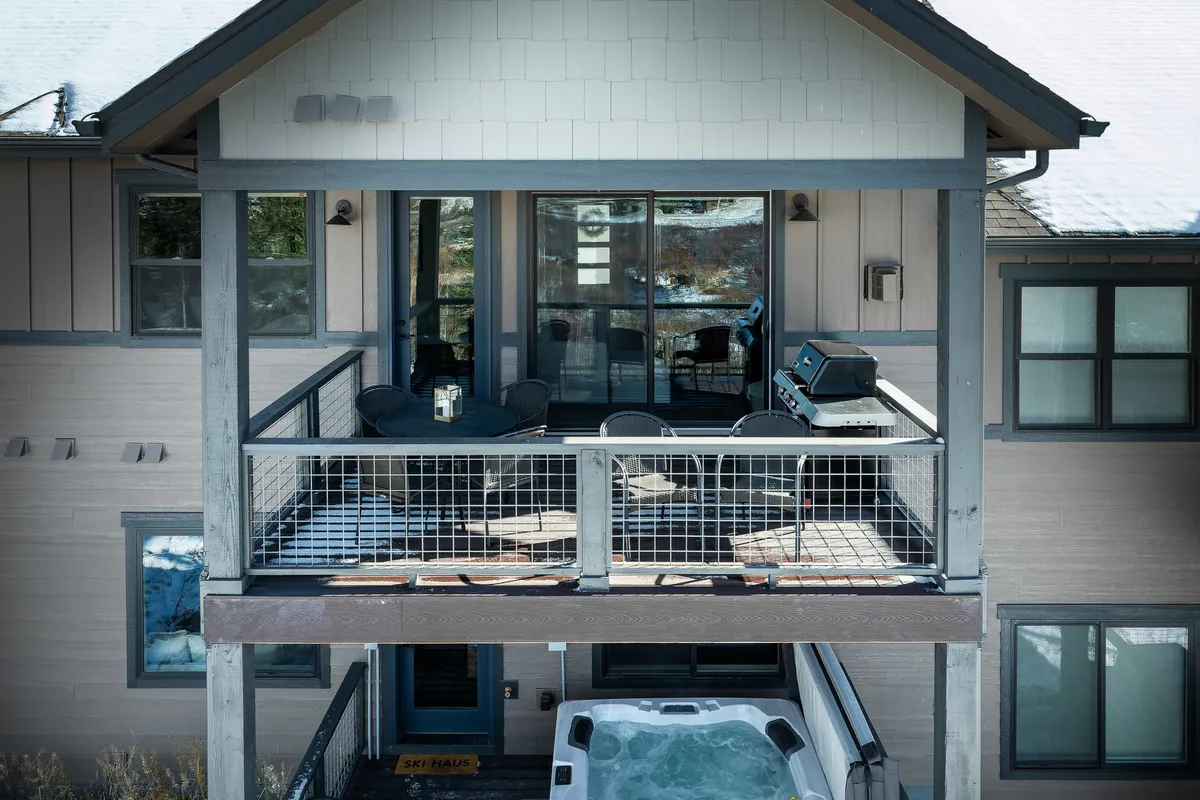 A spacious deck area features a private hot tub surrounded by snow, a grill for outdoor cooking, and inviting seating for relaxation or social gatherings. The backdrop hints at a tranquil and scenic landscape, perfect for unwinding after mountain adventures.
